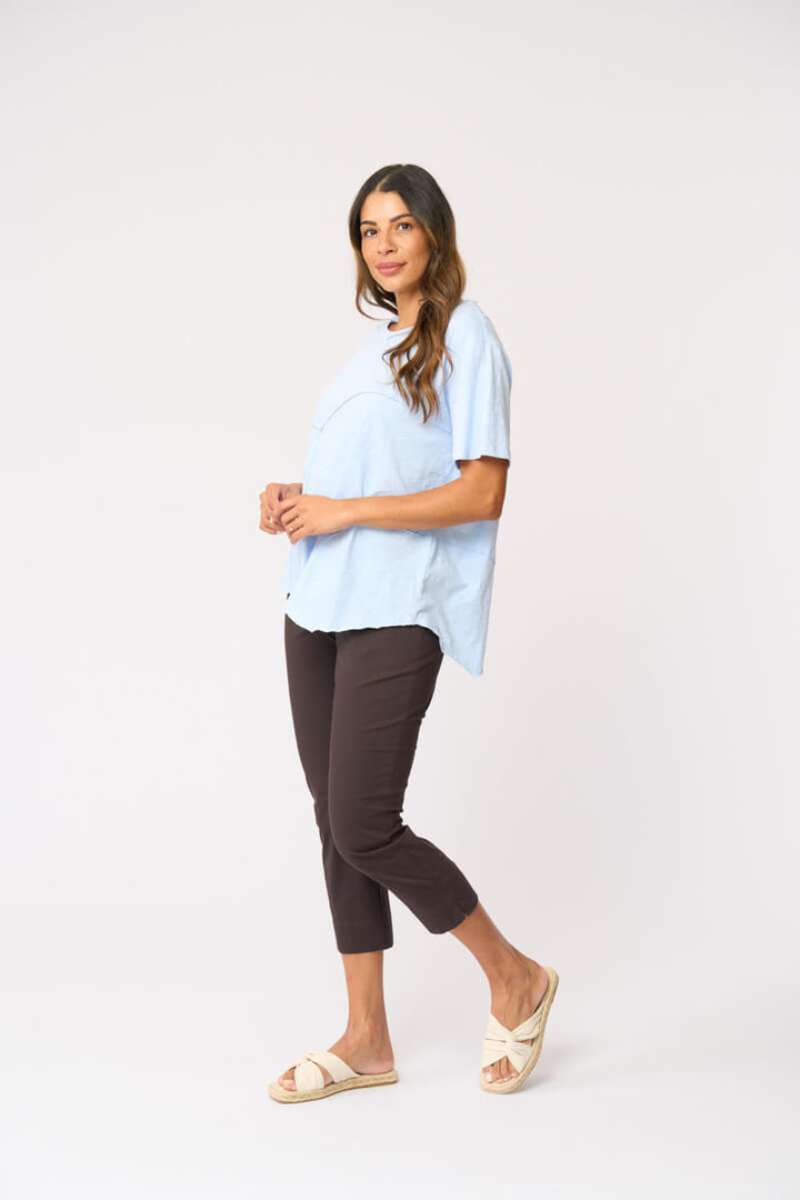 Foil clothing modeling a light blue short-sleeve top with asymmetrical seam details, paired with dark brown cropped pants and beige slide sandals, against a plain white studio background.