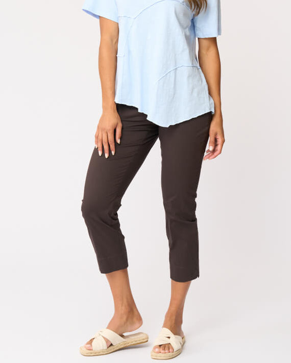 Foil clothing modeling a light blue short-sleeve top with asymmetrical seam details, paired with dark brown cropped pants and beige slide sandals, against a plain white studio background.