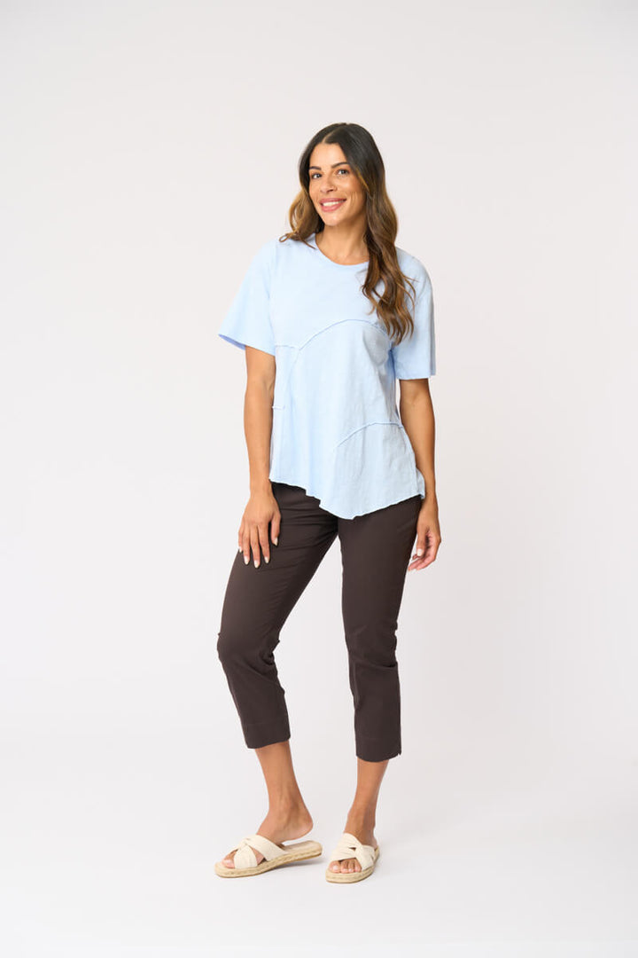 Foil clothing modeling a light blue short-sleeve top with asymmetrical seam details, paired with dark brown cropped pants and beige slide sandals, against a plain white studio background.