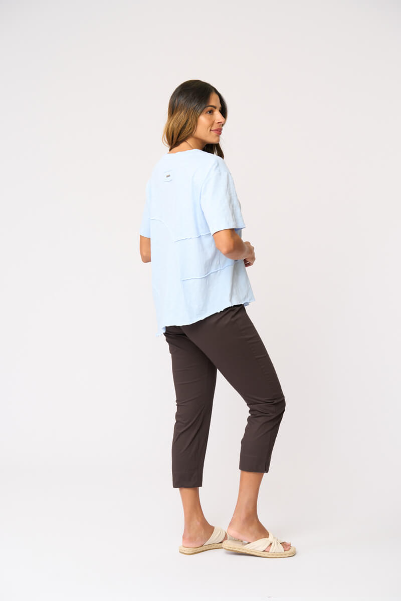 Foil clothing modeling a light blue short-sleeve top with asymmetrical seam details, paired with dark brown cropped pants and beige slide sandals, against a plain white studio background. Back view.