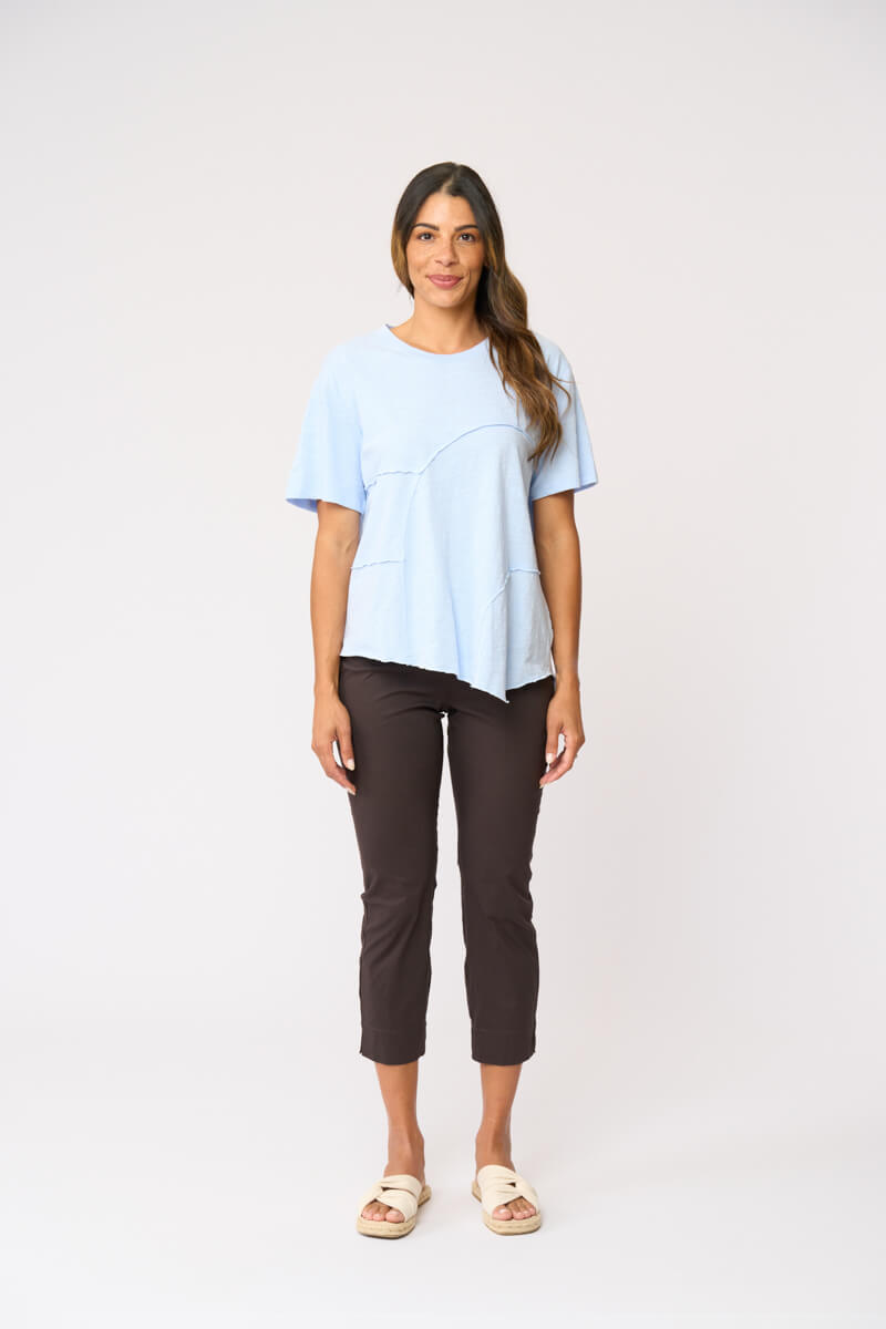 Foil clothing modeling a light blue short-sleeve top with asymmetrical seam details, paired with dark brown cropped pants and beige slide sandals, against a plain white studio background.