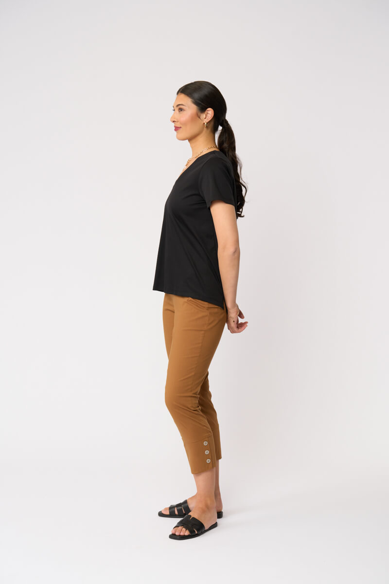 Foil model in a black short-sleeve V-neck t-shirt and cropped tan umber pants with black slide sandals, standing against a plain light gray studio background. Side view