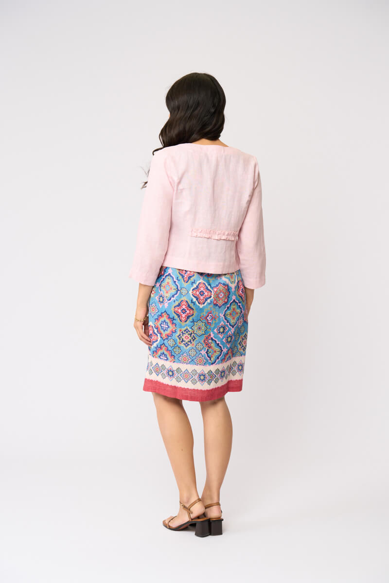 Foil model in a light pink cardigan over a blue and red patterned dress, standing against a plain white studio background. Back view 