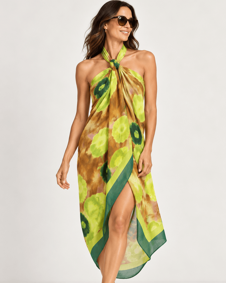 Pom Amsterdam  woman modeling a halter-neck, high-slit wrap dress or sarong in a green and yellow floral print with a brown base, set against a plain light gray studio background.