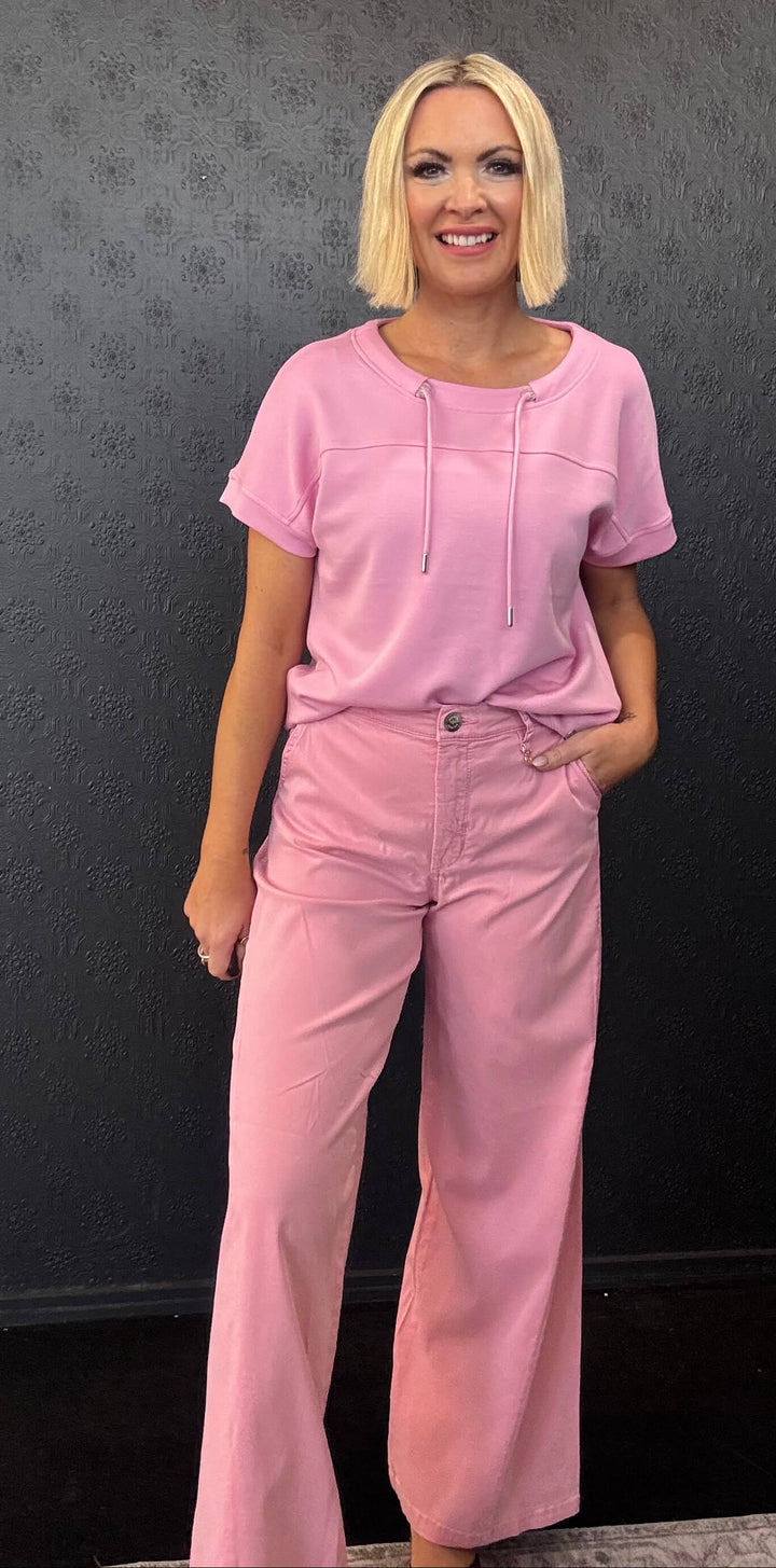 Mos Mosh A model with blonde hair wearing a matching light pink short-sleeve top and wide-leg pants, standing against a dark textured wall.