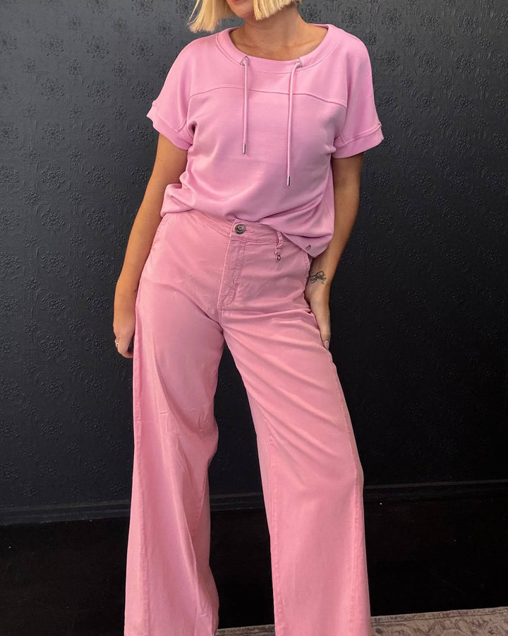 Mos Mosh A model with blonde hair wearing a matching light pink short-sleeve top and wide-leg pants, standing against a dark textured wall.