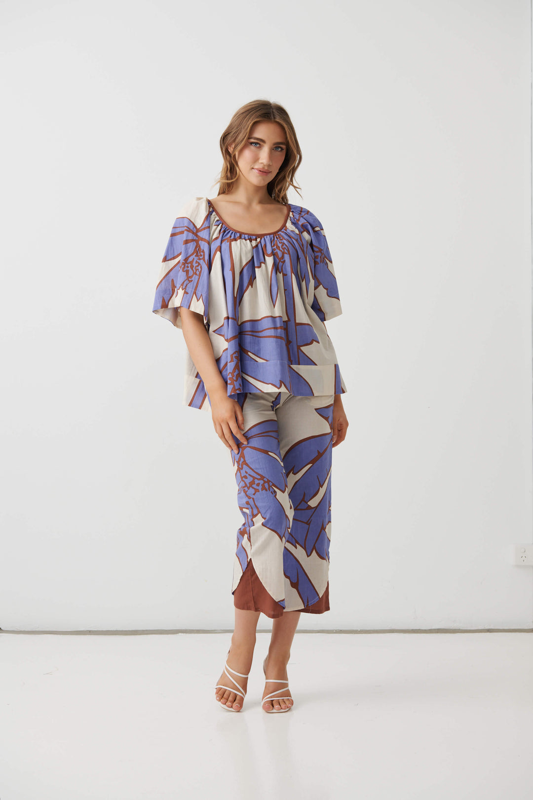 Holmes &amp; Fallon woman modeling a matching two-piece set featuring a short-sleeve top and cropped pants in a blue and brown botanical pattern on a cream background, paired with white heeled sandals. White studio background
