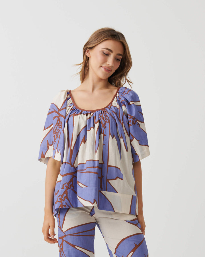 Holmes &amp; Fallon woman modeling a matching two-piece set featuring a short-sleeve top and cropped pants in a blue and brown botanical pattern on a cream background, paired with white heeled sandals. White studio background