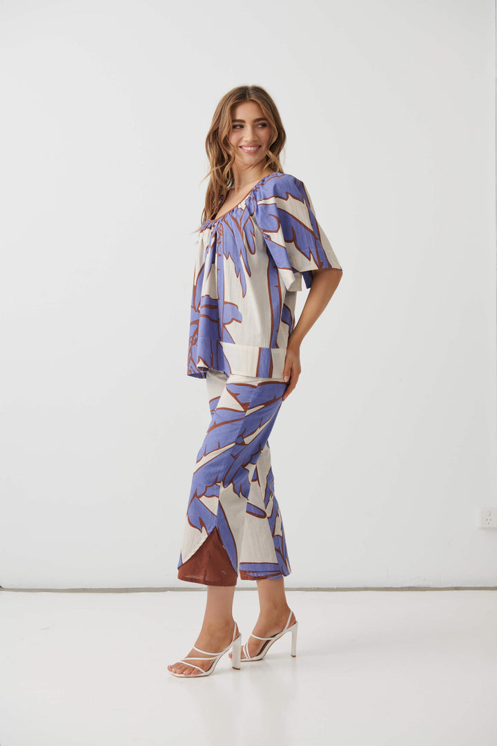 Holmes &amp; Fallon woman modeling a matching two-piece set featuring a short-sleeve top and cropped pants in a blue and brown botanical pattern on a cream background, paired with white heeled sandals. White studio background