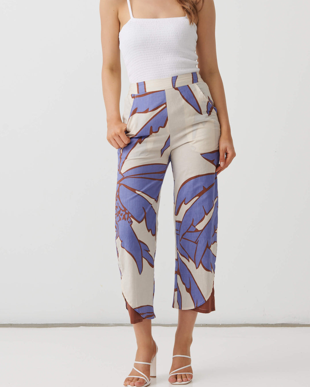 A woman modeling a pair of cropped pants with a blue and purple botanical leaf pattern on a cream background, paired with a white camisole and white strappy heels, set against a plain white studio background.