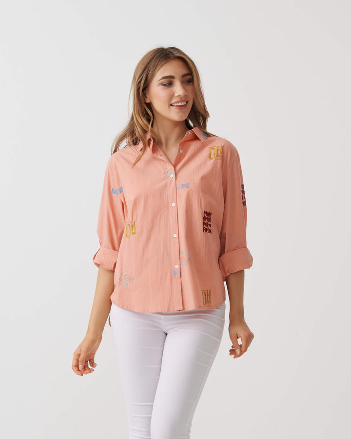 Holmes &amp; Fallon woman modeling a light peach-pink button-up shirt with colorful embroidered details, paired with white capri pants and strappy heels, standing against a plain white studio background.