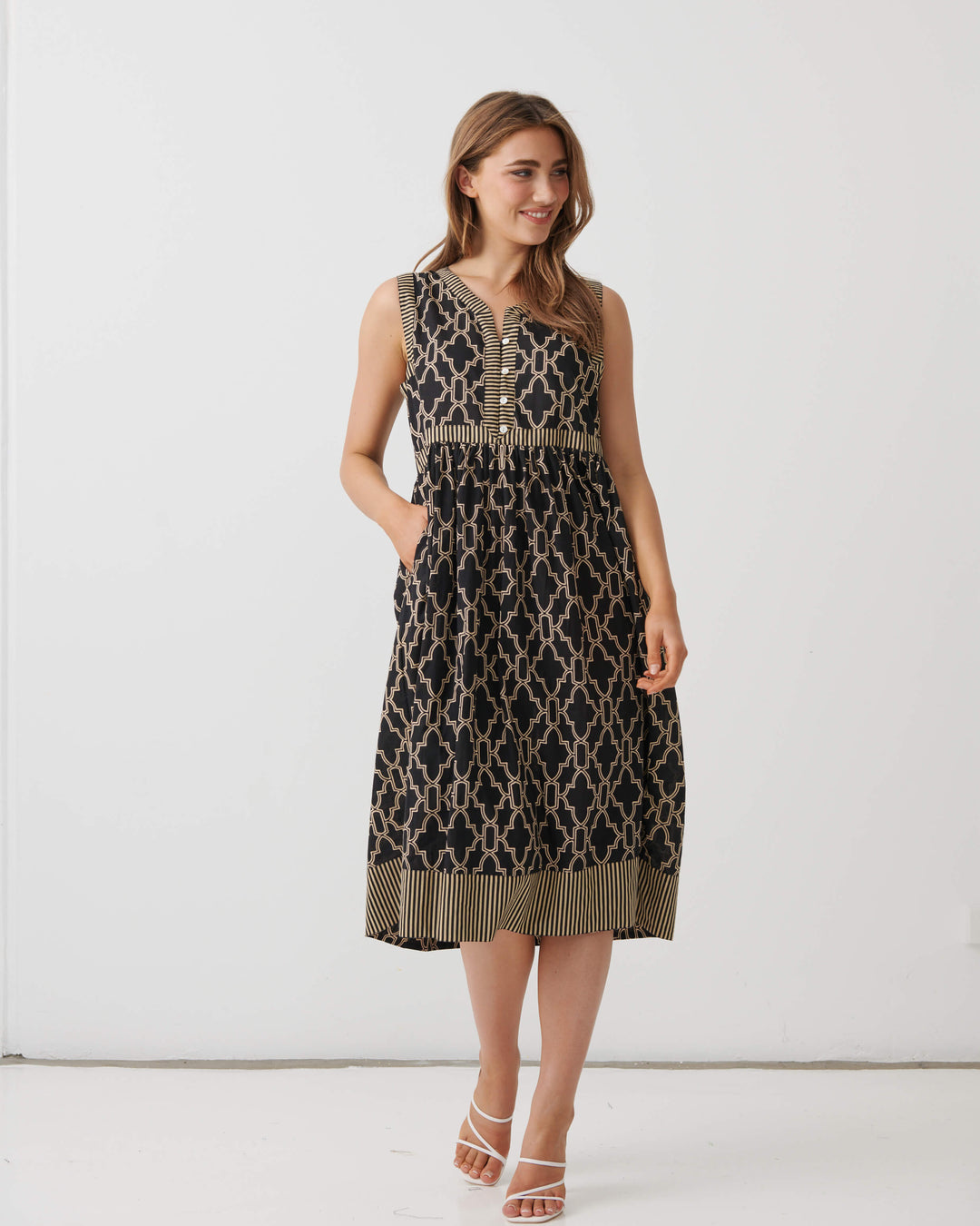 A woman modeling a sleeveless midi dress with a black and beige trellis pattern, featuring a buttoned yoke and a fringed hem, set against a plain white studio background.