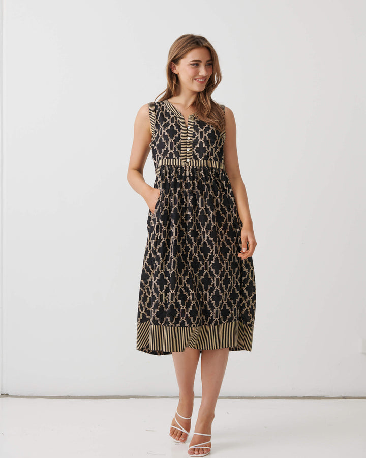 A woman modeling a sleeveless midi dress with a black and beige trellis pattern, featuring a buttoned yoke and a fringed hem, set against a plain white studio background.