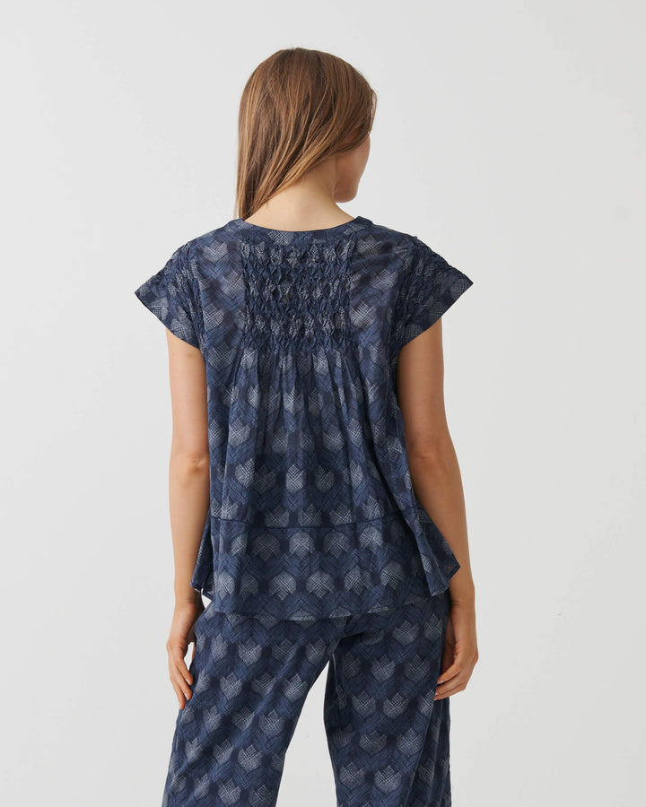 Holmes &amp; Fallon woman modeling a matching two-piece lounge set consisting of a smocked short-sleeve top and wide-leg cropped pants, both in navy blue with a light blue floral pattern, set against a plain white studio background.. Back view