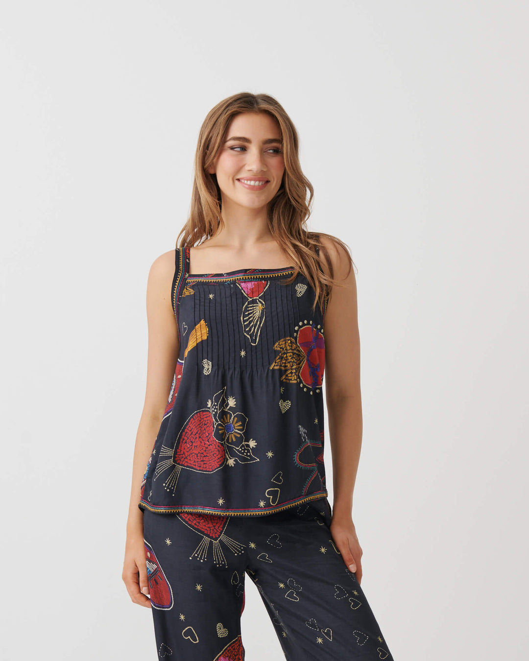 A woman modeling a matching two-piece lounge set consisting of a sleeveless top and wide-leg pants in navy blue with a colorful abstract print, paired with white high-heeled sandals, set against a plain white studio background.