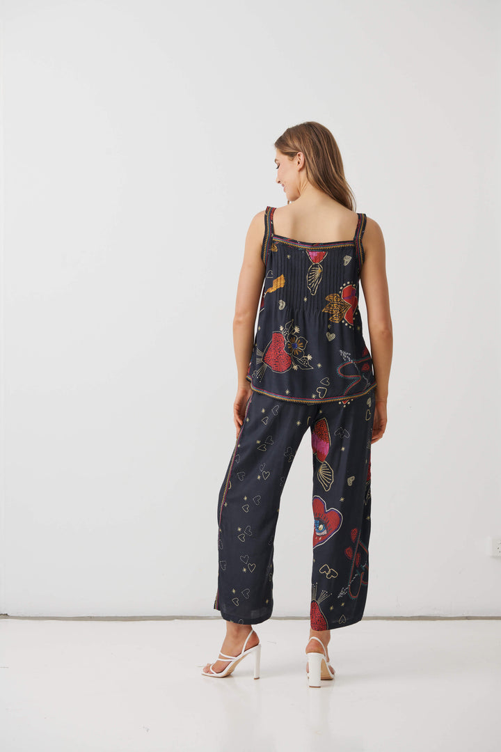 A woman modeling a matching two-piece lounge set consisting of a sleeveless top and wide-leg pants in navy blue with a colorful abstract print, paired with white high-heeled sandals, set against a plain white studio background. back view 