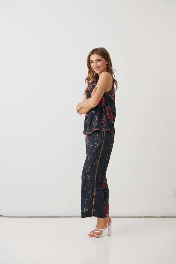A woman modeling a matching two-piece lounge set consisting of a sleeveless top and wide-leg pants in navy blue with a colorful abstract print, paired with white high-heeled sandals, set against a plain white studio background.