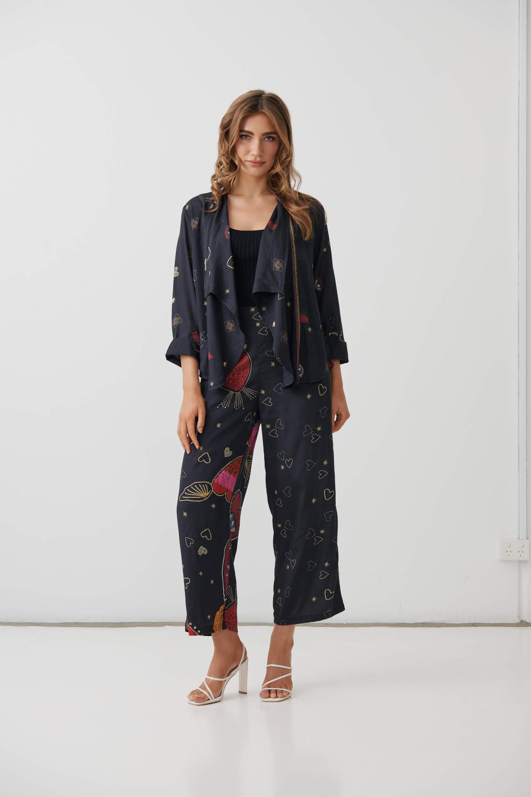 Holmes &amp; Fallon  woman modeling a dark navy two-piece lounge set with a colorful abstract print, consisting of a short-sleeve top and matching wide-leg pants, paired with a black tank top and beige strappy heels. On a white studio background
