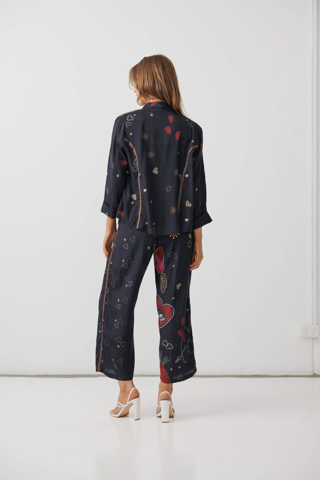 Holmes &amp; Fallon  woman modeling a dark navy two-piece lounge set with a colorful abstract print, consisting of a short-sleeve top and matching wide-leg pants, paired with a black tank top and beige strappy heels. On a white studio background. Back view