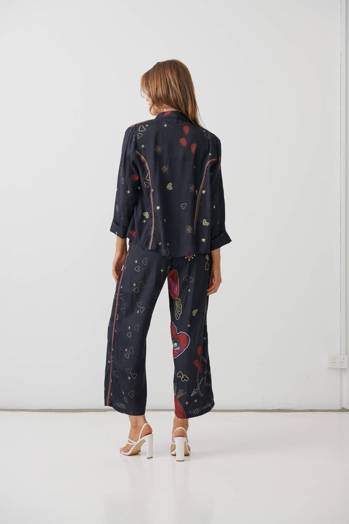 Holmes &amp; Fallon  woman modeling a dark navy two-piece lounge set with a colorful abstract print, consisting of a short-sleeve top and matching wide-leg pants, paired with a black tank top and beige strappy heels. On a white studio background. Back view