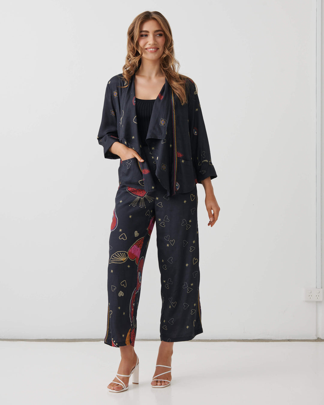 Holmes &amp; Fallon model wearing high-waisted, wide-leg cropped pants in a dark navy blue base with a colorful graphic print of red hearts, gold hearts, and small gold stars, paired with a black ribbed tank top and white heeled sandals.
