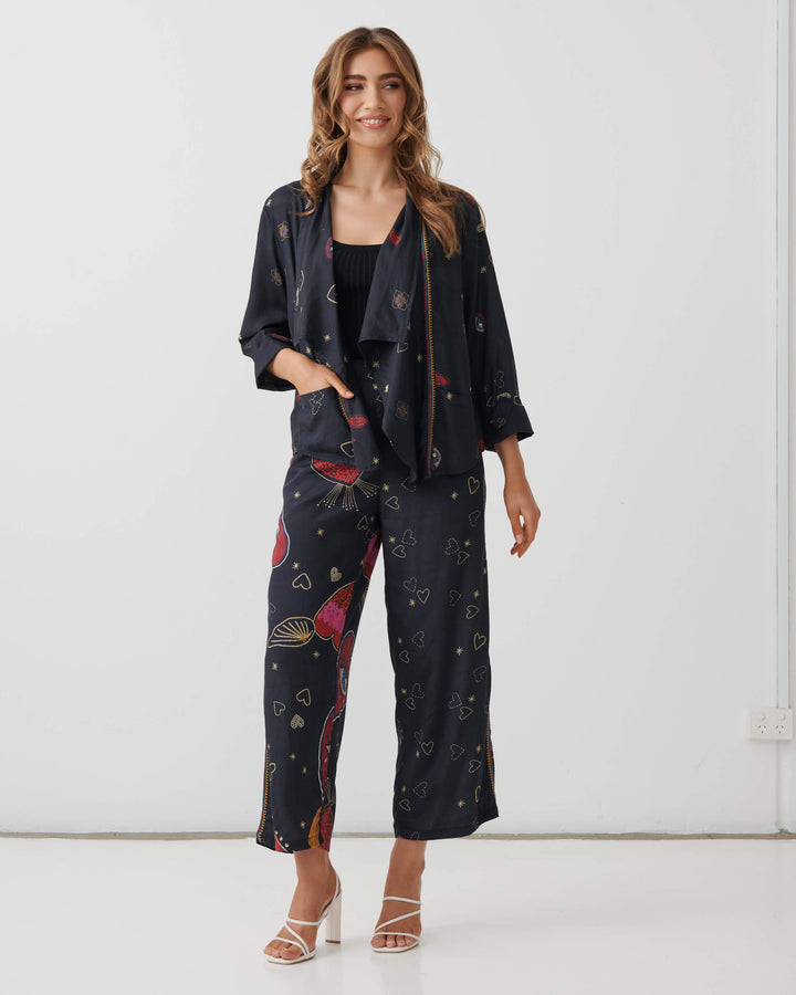 Holmes &amp; Fallon model wearing high-waisted, wide-leg cropped pants in a dark navy blue base with a colorful graphic print of red hearts, gold hearts, and small gold stars, paired with a black ribbed tank top and white heeled sandals.