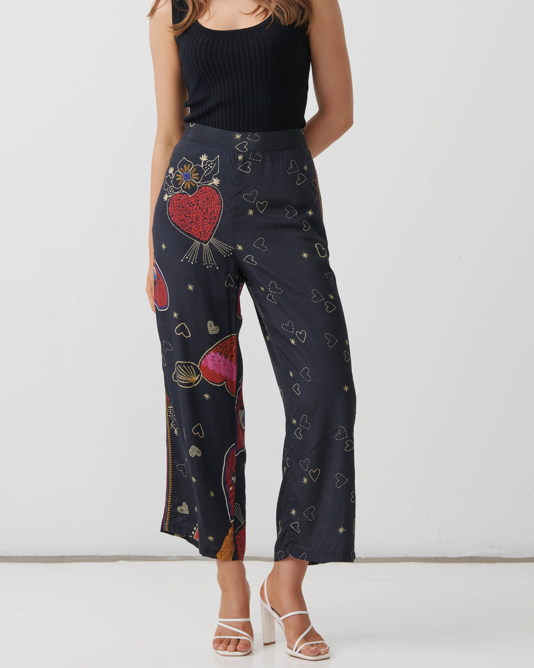 Holmes &amp; Fallon model wearing high-waisted, wide-leg cropped pants in a dark navy blue base with a colorful graphic print of red hearts, gold hearts, and small gold stars, paired with a black ribbed tank top and white heeled sandals.