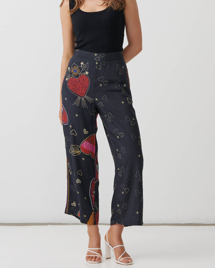 Holmes &amp; Fallon model wearing high-waisted, wide-leg cropped pants in a dark navy blue base with a colorful graphic print of red hearts, gold hearts, and small gold stars, paired with a black ribbed tank top and white heeled sandals.