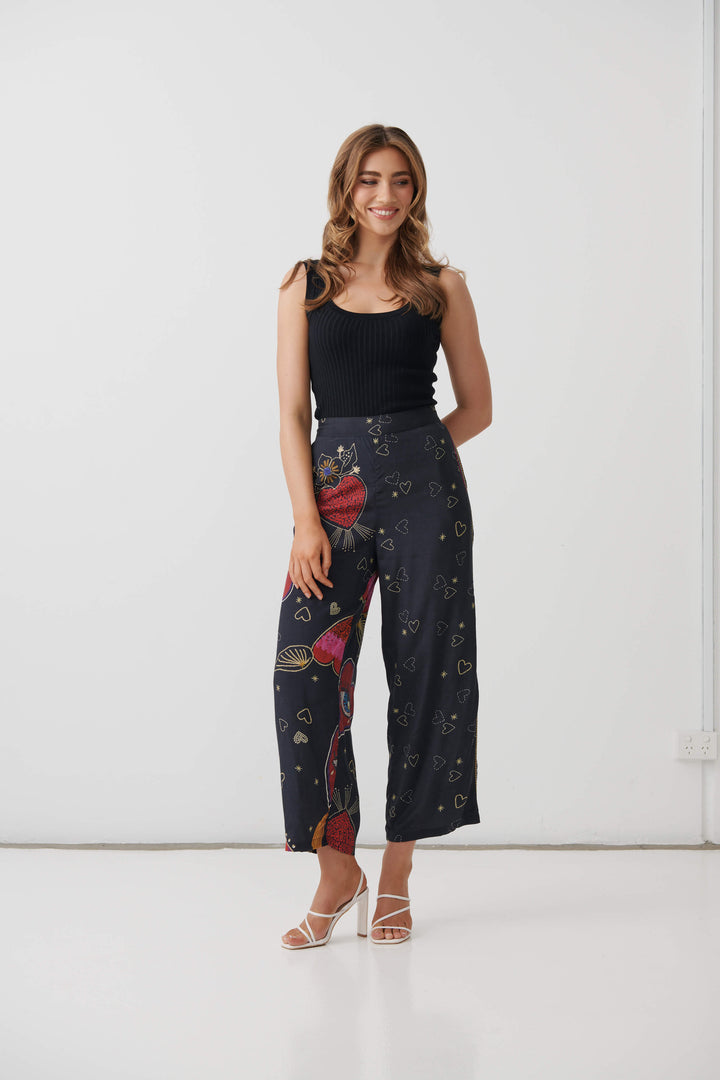 Holmes &amp; Fallon model wearing high-waisted, wide-leg cropped pants in a dark navy blue base with a colorful graphic print of red hearts, gold hearts, and small gold stars, paired with a black ribbed tank top and white heeled sandals.