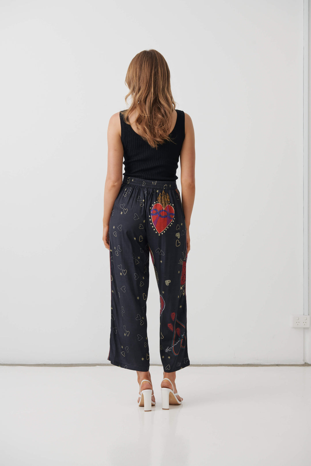 Holmes &amp; Fallon model wearing high-waisted, wide-leg cropped pants in a dark navy blue base with a colorful graphic print of red hearts, gold hearts, and small gold stars, paired with a black ribbed tank top and white heeled sandals. Back view 