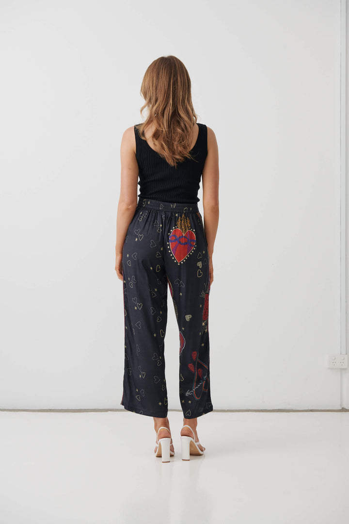 Holmes &amp; Fallon model wearing high-waisted, wide-leg cropped pants in a dark navy blue base with a colorful graphic print of red hearts, gold hearts, and small gold stars, paired with a black ribbed tank top and white heeled sandals. Back view 