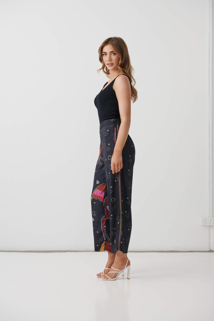 Holmes &amp; Fallon model wearing high-waisted, wide-leg cropped pants in a dark navy blue base with a colorful graphic print of red hearts, gold hearts, and small gold stars, paired with a black ribbed tank top and white heeled sandals.