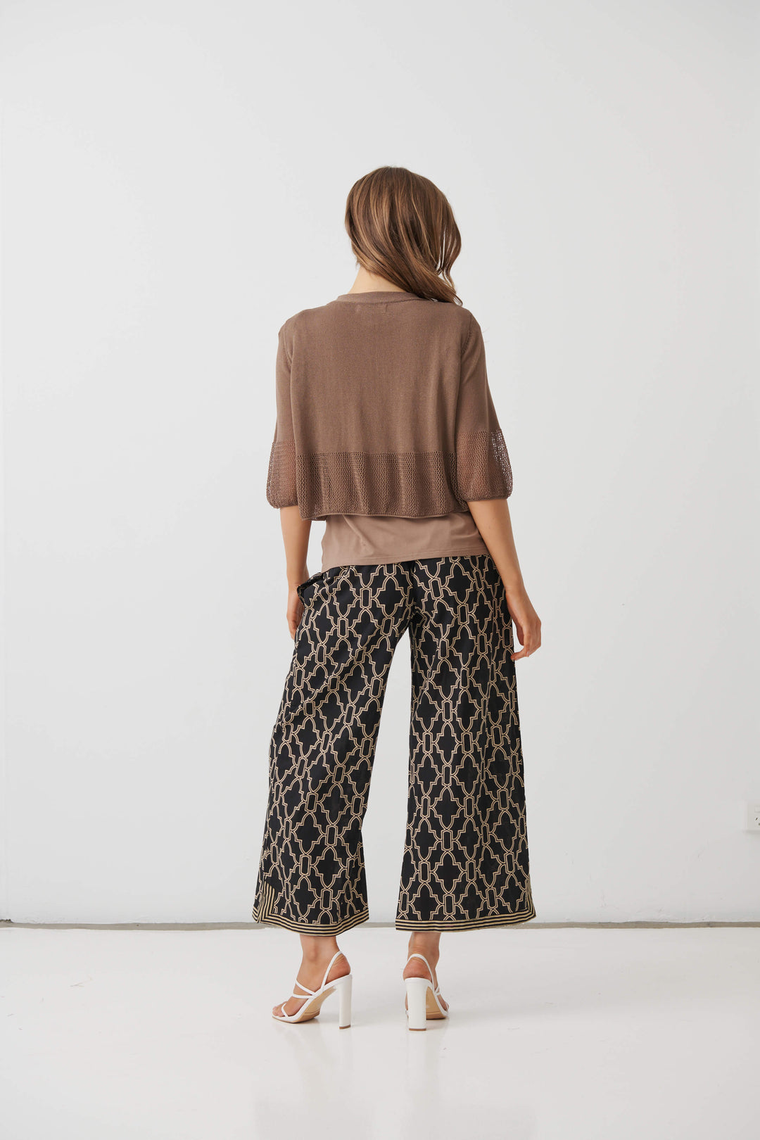 Holmes &amp; Fallon  modeling a brown textured three-quarter sleeve top paired with black wide-leg pants featuring a beige trellis pattern, standing against a plain white studio background. Back view 
