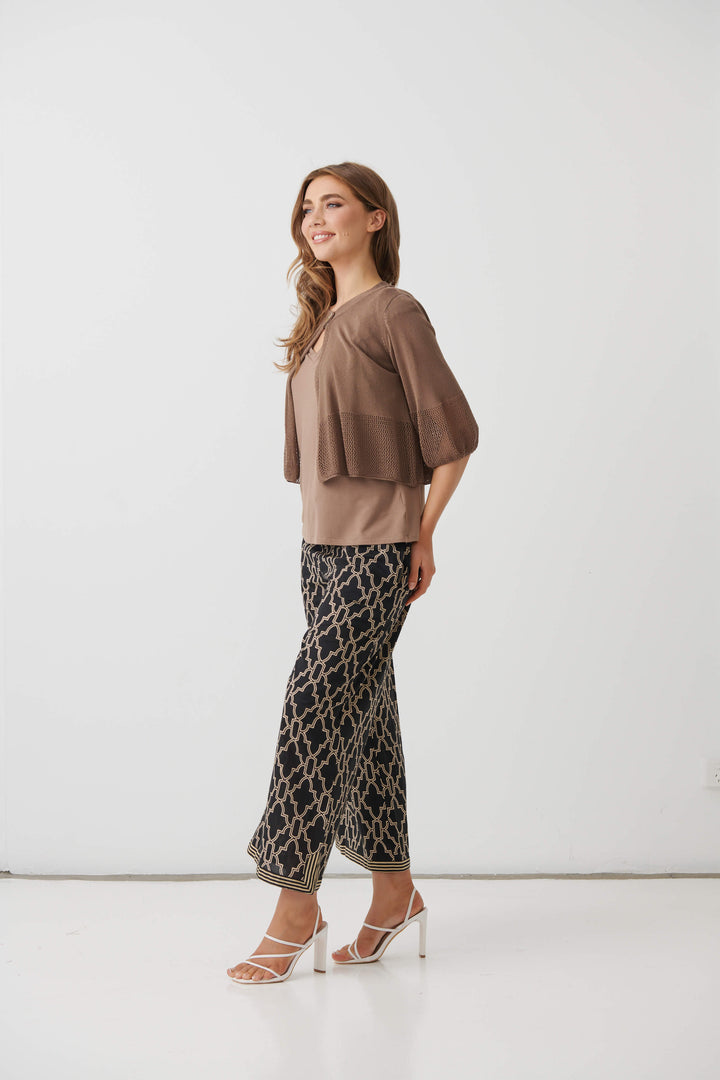 Holmes &amp; Fallon  modeling a brown textured three-quarter sleeve top paired with black wide-leg pants featuring a beige trellis pattern, standing against a plain white studio background.