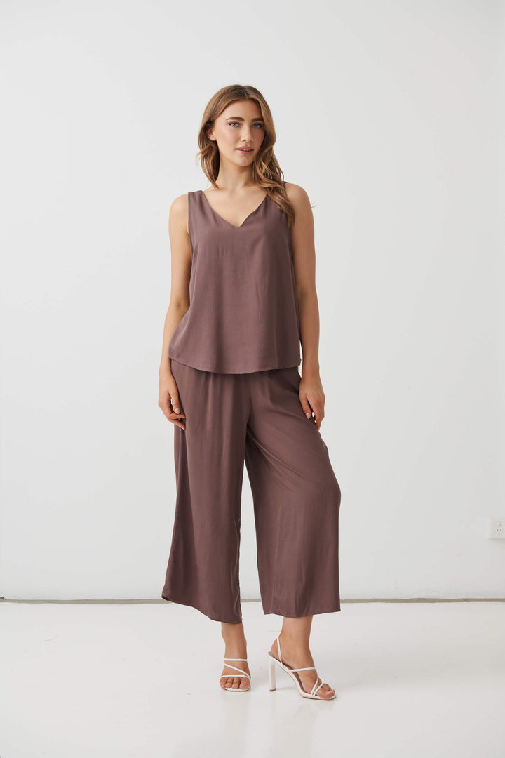 Holmes &amp; Fallon  modeling a sleeveless brown wide-leg pant with a drawstring waist, paired with white strappy high-heeled sandals, against a plain white studio background.
