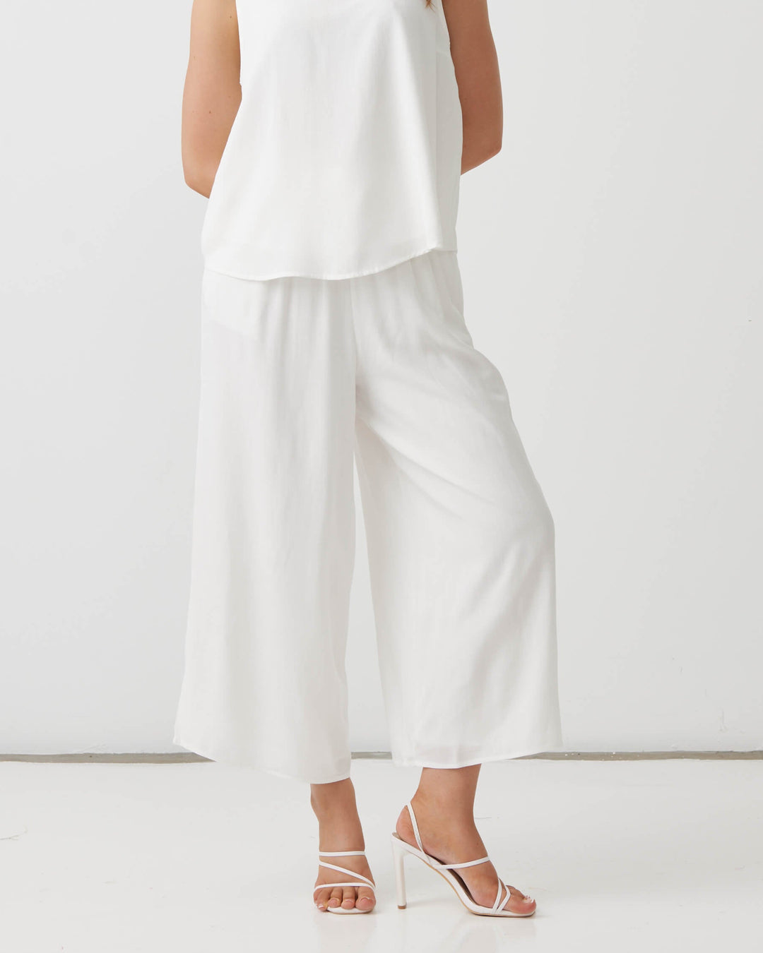 Holmes &amp; Fallon modeling a white sleeveless V-neck top and matching white wide-leg cropped pants, paired with white strappy high-heeled sandals, against a plain white studio background.