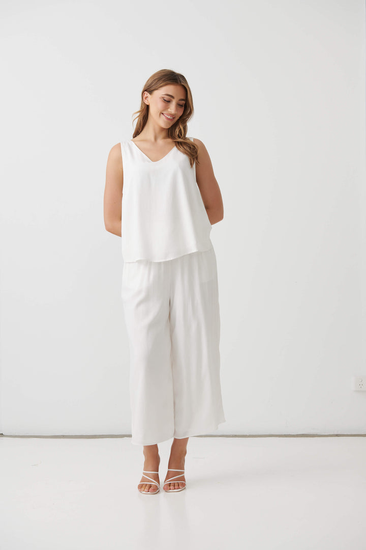 Holmes &amp; Fallon modeling a white sleeveless V-neck top and matching white wide-leg cropped pants, paired with white strappy high-heeled sandals, against a plain white studio background.