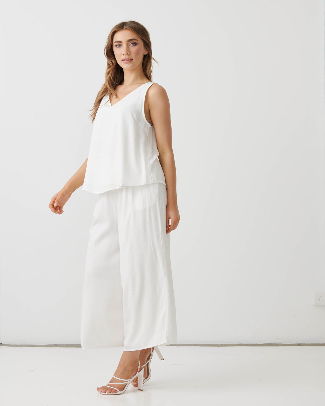 Holmes &amp; Fallon modeling a white sleeveless V-neck top and matching white wide-leg cropped pants, paired with white strappy high-heeled sandals, against a plain white studio background.