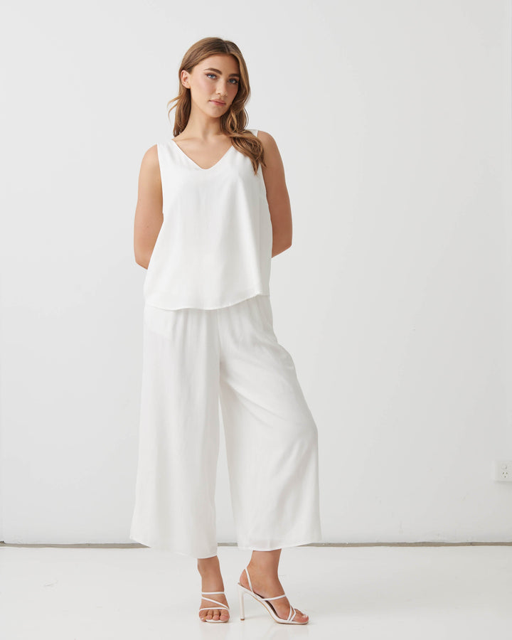 Holmes &amp; Fallon modeling a white sleeveless V-neck top and matching white wide-leg cropped pants, paired with white strappy high-heeled sandals, against a plain white studio background.