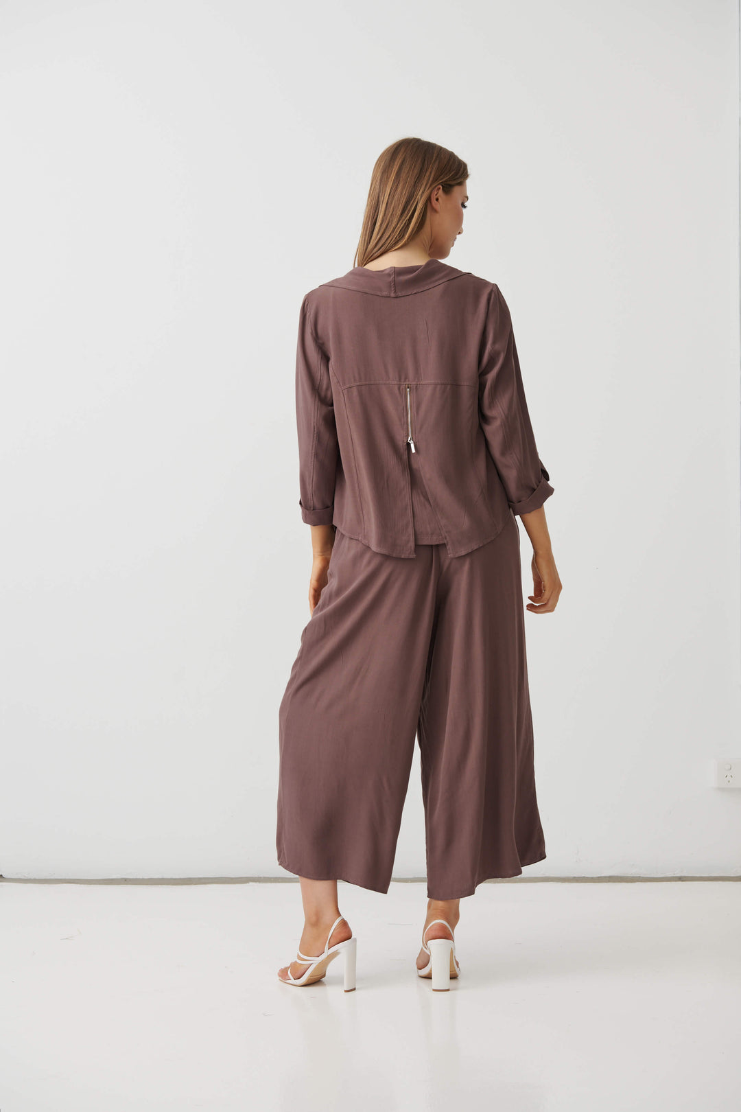 Holmes &amp; Fallon  modeling a sleeveless brown wide-leg pant with a drawstring waist, paired with white strappy high-heeled sandals, against a plain white studio background.