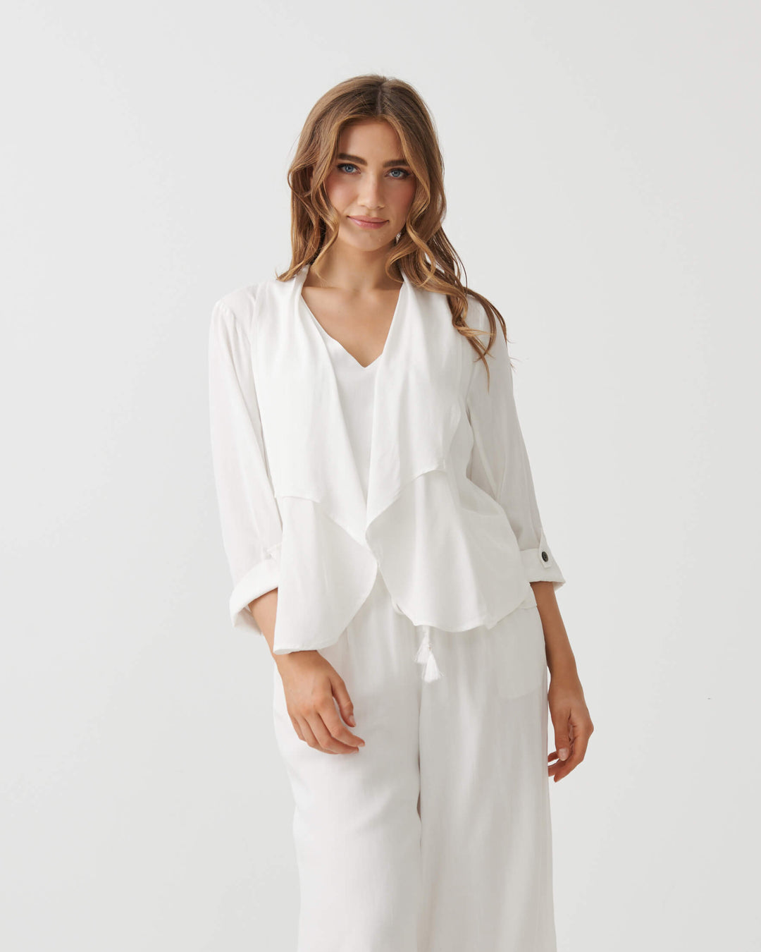 Holmes &amp; Fallon modeling a white two-piece outfit consisting of a draped long-sleeve blouse and matching wide-leg cropped pants, paired with strappy high-heeled sandals against a plain white studio background.