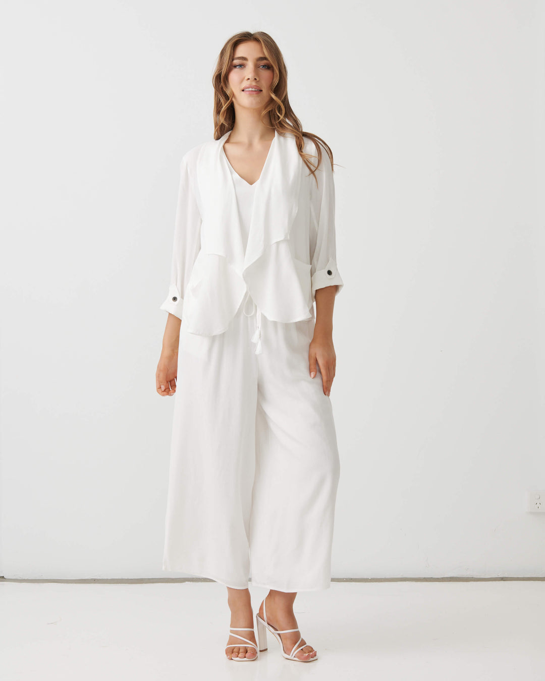 Holmes &amp; Fallon modeling a white two-piece outfit consisting of a draped long-sleeve blouse and matching wide-leg cropped pants, paired with strappy high-heeled sandals against a plain white studio background.