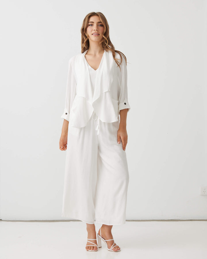 Holmes &amp; Fallon modeling a white two-piece outfit consisting of a draped long-sleeve blouse and matching wide-leg cropped pants, paired with strappy high-heeled sandals against a plain white studio background.