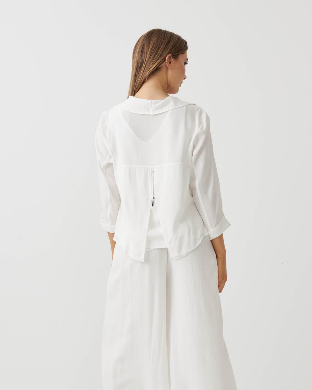 Holmes &amp; Fallon modeling a white two-piece outfit consisting of a draped long-sleeve blouse and matching wide-leg cropped pants, paired with strappy high-heeled sandals against a plain white studio background. back view