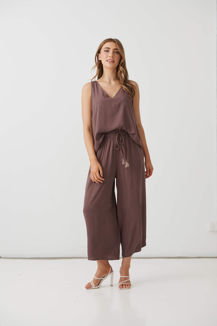 Holmes &amp; Fallon  modeling a sleeveless brown wide-leg pant with a drawstring waist, paired with white strappy high-heeled sandals, against a plain white studio background.