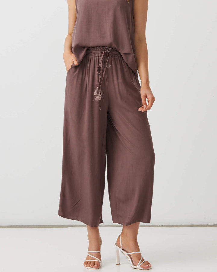 Holmes &amp; Fallon  modeling a sleeveless brown wide-leg pant with a drawstring waist, paired with white strappy high-heeled sandals, against a plain white studio background.