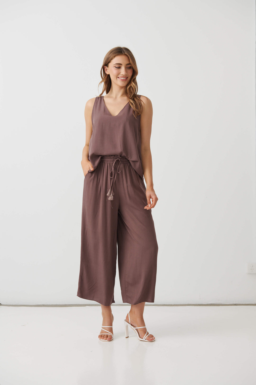 Holmes &amp; Fallon  modeling a sleeveless brown wide-leg pant with a drawstring waist, paired with white strappy high-heeled sandals, against a plain white studio background.
