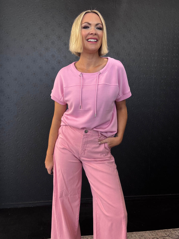 Mos Mosh A model with blonde hair wearing a matching light pink short-sleeve top and wide-leg pants, standing against a dark textured wall.