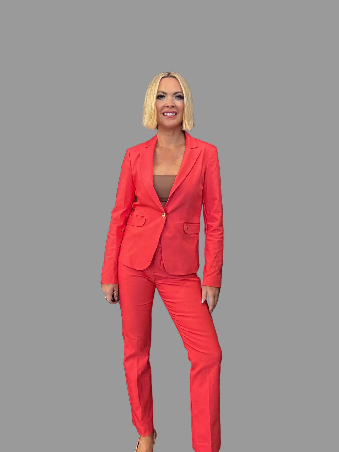 Mos Mosh A model with a blonde bob wearing a bright coral pink tailored pantsuit over a brown top, posing against a dark textured wall.