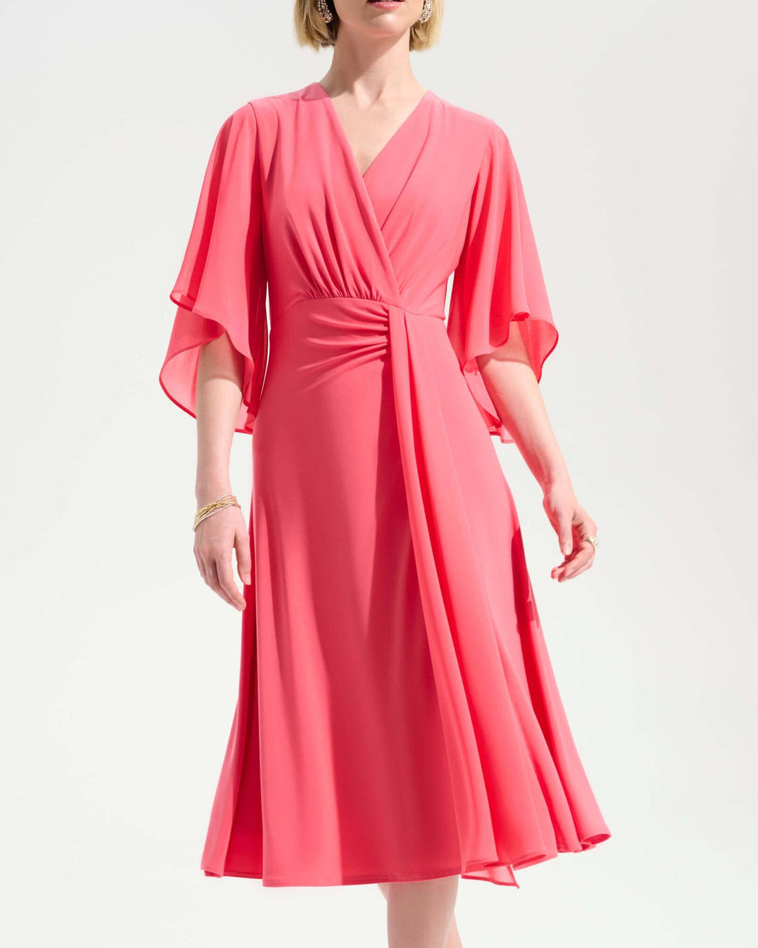 Joseph Ribkoff modelling a Coral pink fit-and-flare sash dress with V-neck and flutter sleeves, midi length. On a pale studio background